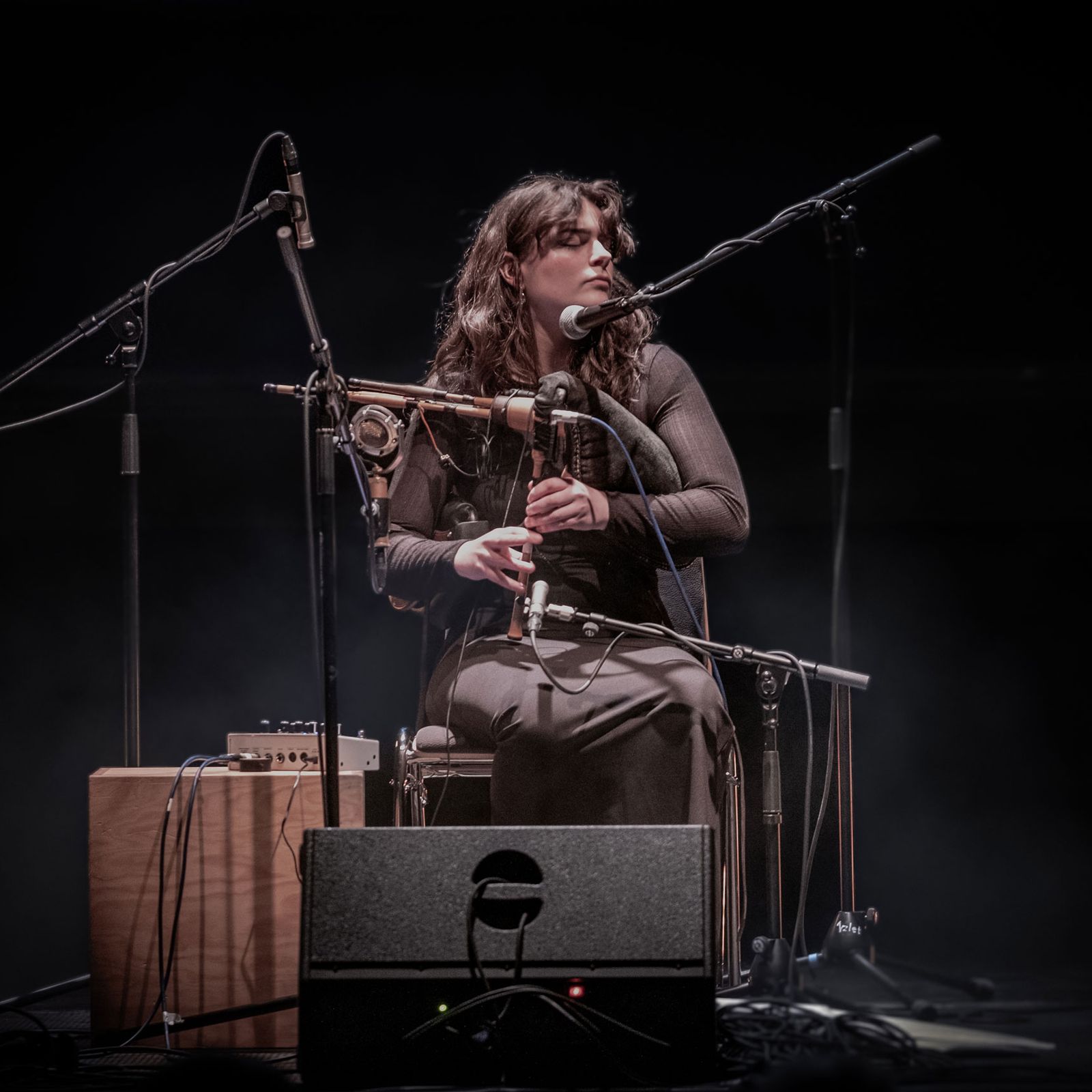 A woman on a dark stage playing the bagpipes with microphones around her.
