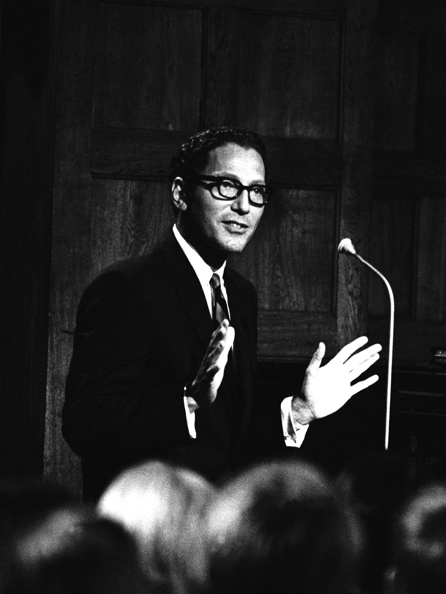 Figure in glasses suit and tie stands in front of microphone looking out into audience with hands in an open gesture.