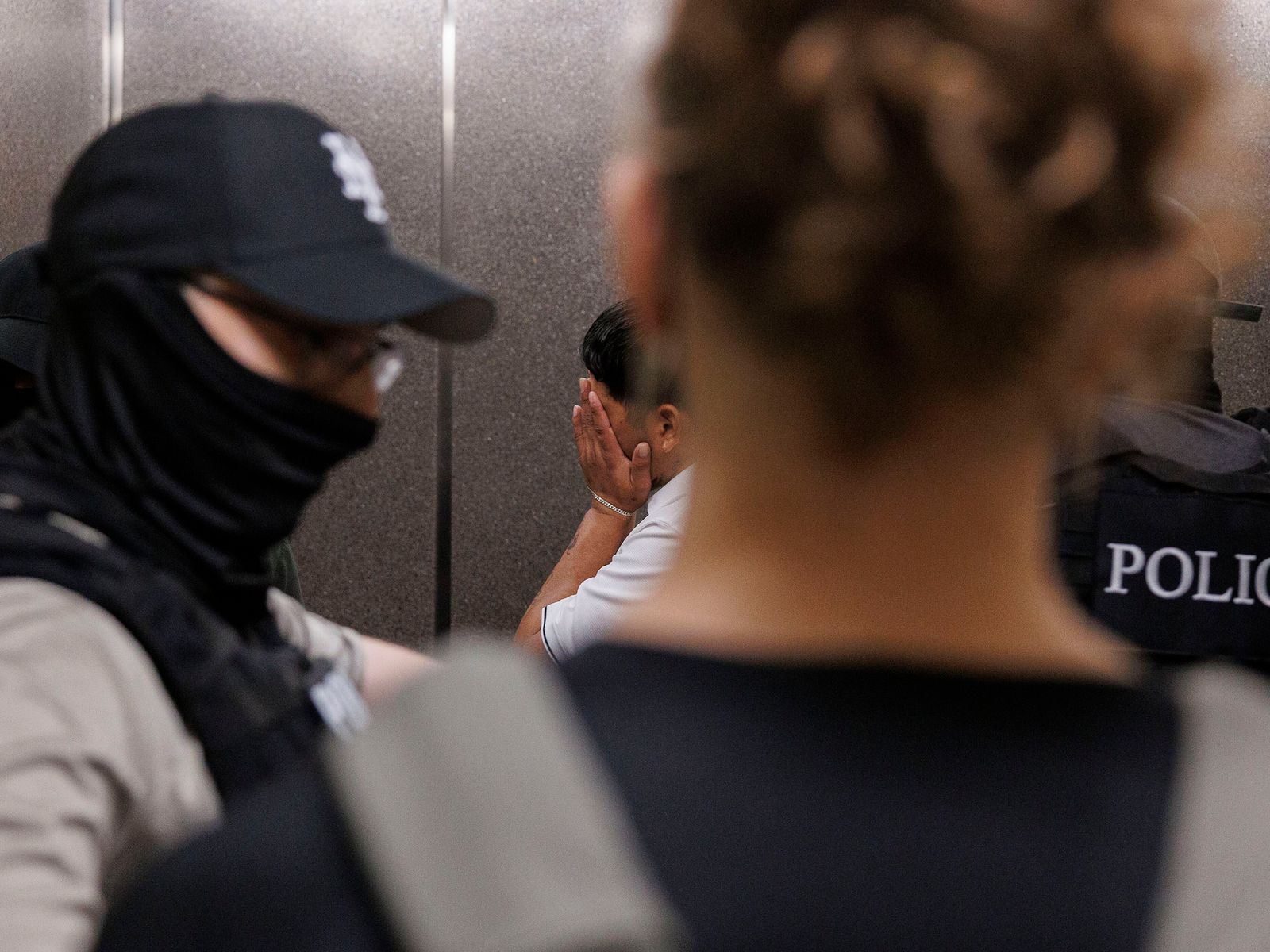 A person stands in distance with face covered by hands while ICE officers and police stand close to viewer.